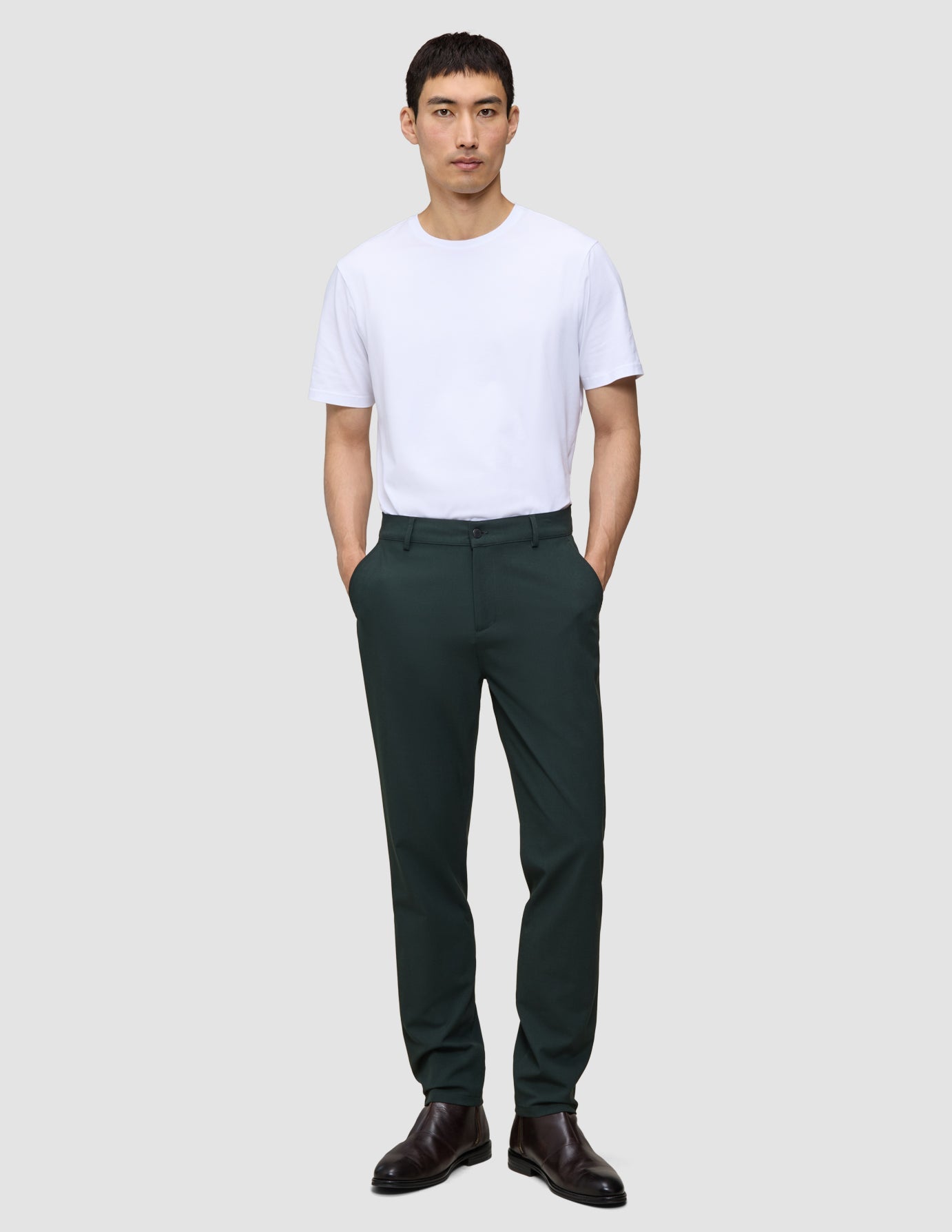 En mann i en hvit T-skjorte og Ease Pants Regular Northern Pine står med hendene i lommene.